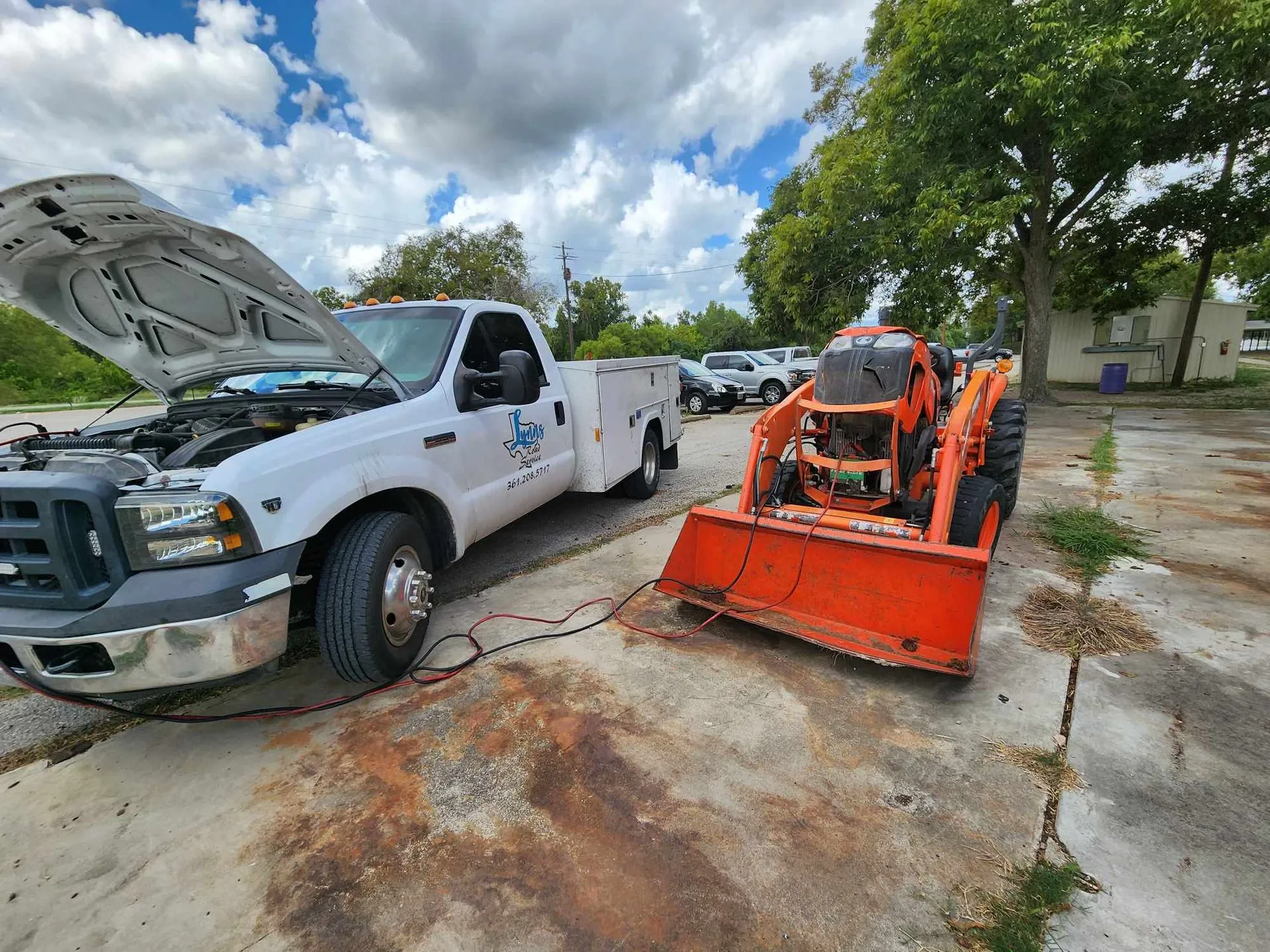 Service truck jump-starting an orange skid steer in a paved lot.