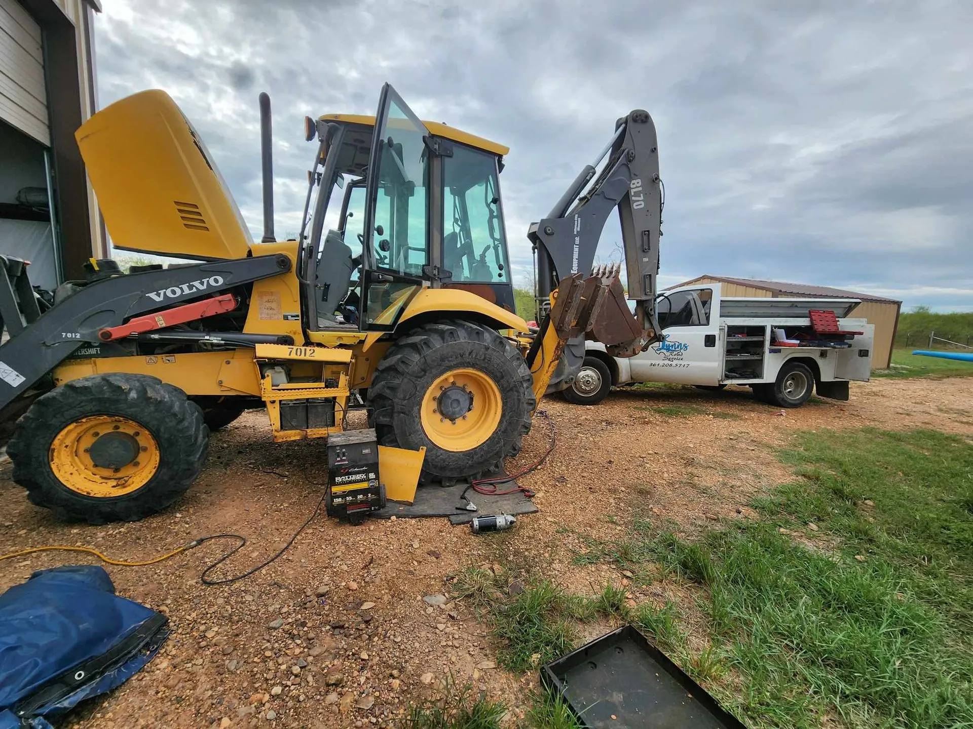 Yellow backhoe with its hood up parked near a service truck.