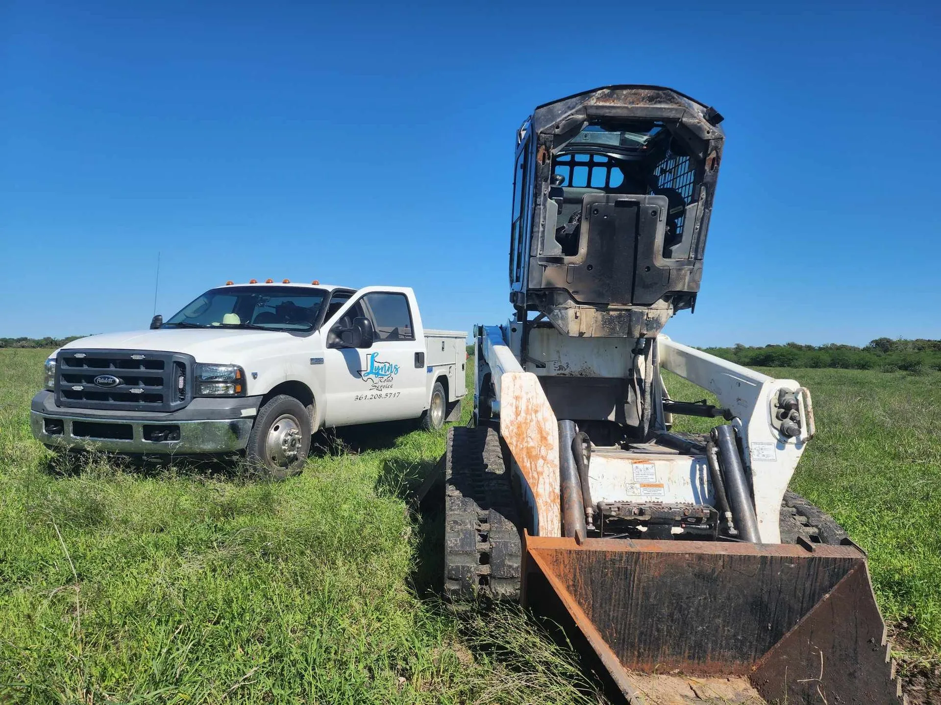 A white pickup truck and a skid steer in a grassy field under a blue sky.