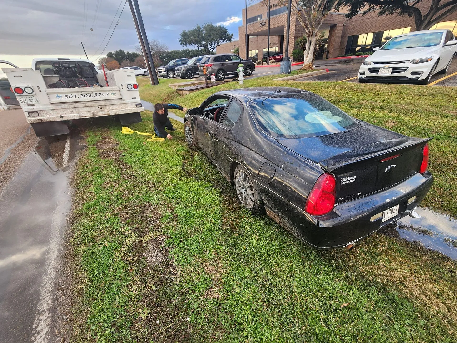 Black car stuck in a ditch with a worker assisting and a tow truck nearby.